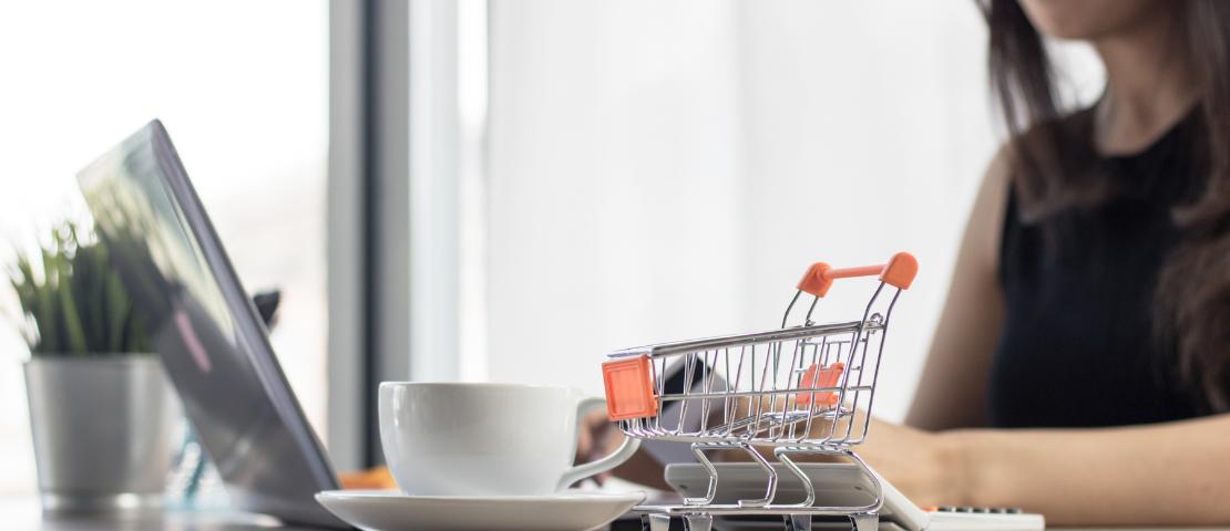 Mulher usando notebook. Sob a mesa, um carrinho de compras em miniatura com detalhes em laranja e uma xícara de café, simbolizando organização de catálogo na Shopee para vendedores online.